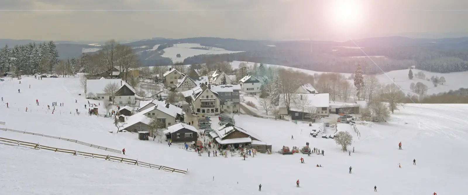 Skiërs op een brede helling boven een besneeuwd dorp, met beboste heuvels tot aan de horizon